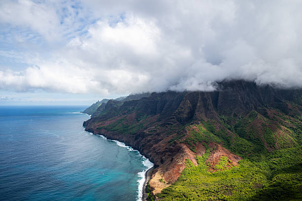 An aerial view of the Na Pali Coast on Kauai, Hawaii, where steep volcanic cliffs meet the turquoise waters of the Pacific Ocean. Low clouds sweep across the ridgelines, softening the dramatic landscape below. The mixture of red earth, green vegetation, and deep blue sea creates a striking natural contrast.