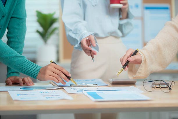 istock Three Asian women collaborate at shared office desk filled with documents, charts, coffee. One leads the discussion, another points at data, third listens attentively, showing productive teamwork. 2239681875
