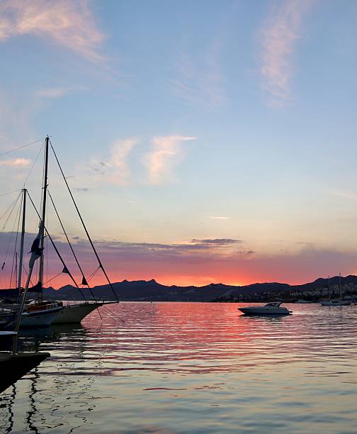 A tranquil harbor at sunset with colorful clouds and a vibrant red-orange sky reflecting on the calm water, framed by silhouettes of docked sailboats. High quality photo