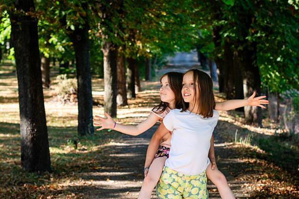 Two young girls having fun together, one giving the other a piggyback ride in a sunny park with trees