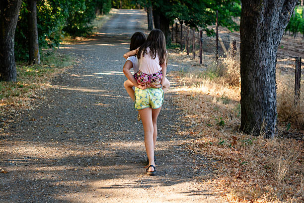 Two young sisters enjoying a piggyback ride on a sunny rural path, representing childhood fun, bond, and support