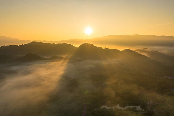 Breathtaking high-angle drone shot capturing the ethereal moment the rising sun pierces through a thick blanket of fog, illuminating the mountain in a stunning golden haze. An incredible view from above the clouds.