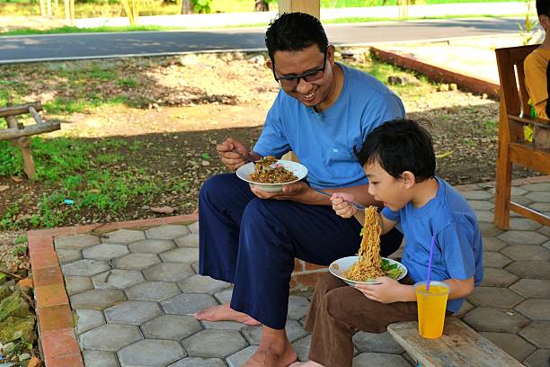 Asian father teaching his young son to eat outdoors during family lunch in countryside atmosphere.