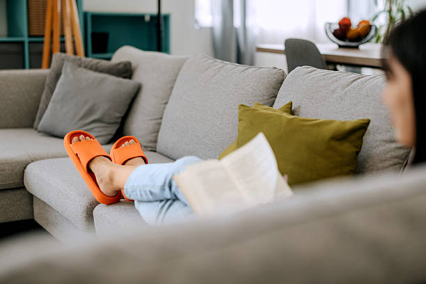 Relaxed woman wearing comfortable rubber slippers while lying on a sofa and reading a book at home. Represents leisure, comfort, and peaceful moments of everyday life.