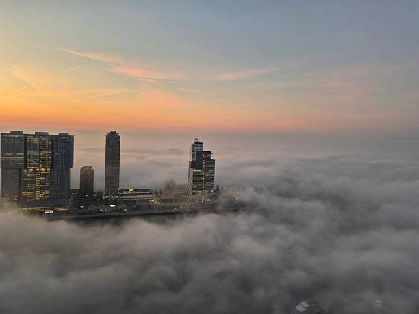Downtown Rotterdam skyline over river Meuse seen from high angle with blanket of thick fog or mist covering everything except the highest tips of the Erasmus bridge and top of skyscrapers. Sunrise creates pink color in the sky. Winter phenomenon in harbor city