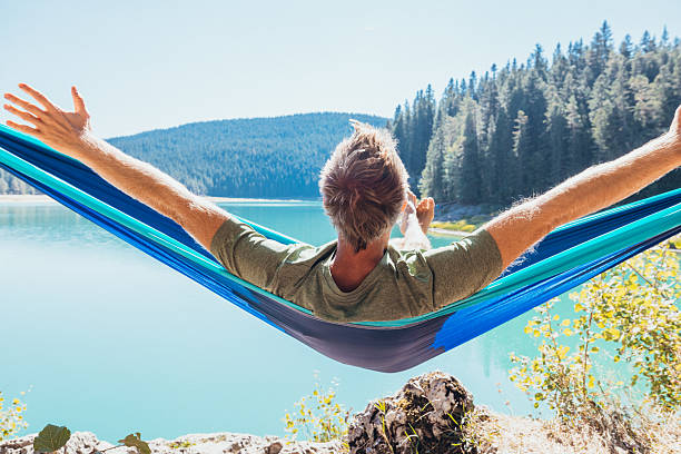 Man with outstretched arms lying in blue hammock overlooking calm lake surrounded by pine trees. Peaceful outdoor relaxation in nature. Scenic forest and water view.
