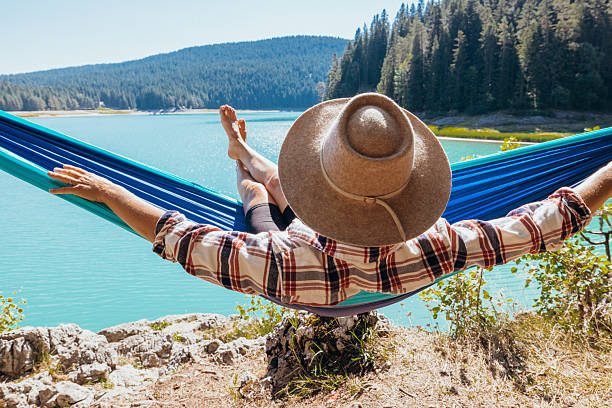 Woman wearing hat and plaid shirt resting in blue hammock near clear lake with forested hills. Peaceful outdoor scene with nature and water. Relaxation and leisure in natural setting.