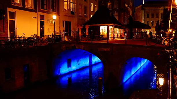 A stunning nighttime view of an Amsterdam canal, showcasing a vibrant blue reflection in the water. The scene is enhanced by warm light from the surrounding buildings and the presence of traditional bicycles.