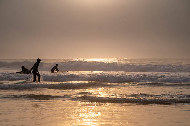 Surfers prepare to ride the waves during a warm, glowing sunset in Colares, Portugal, capturing the spirit of adventure and the beauty of the ocean.