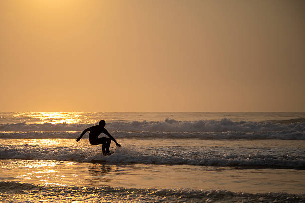A surfer balances on a wave during golden sunset in Colares, Portugal, with shimmering reflections on the Atlantic Ocean creating a peaceful coastal scene.