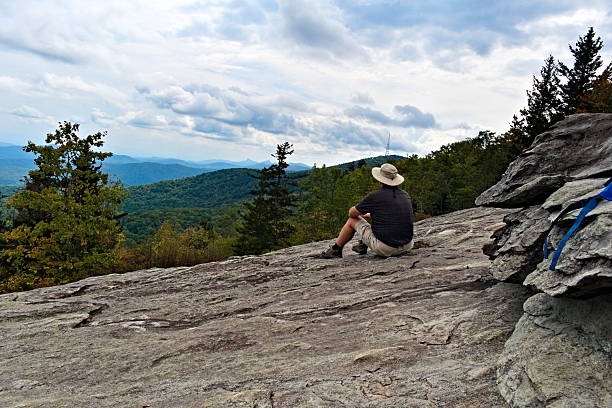 A solo hiker, in the Blue Ridge Mountains, sits on a rocky overlook and enjoys looking out over the valley after hiking to the top of a trail.