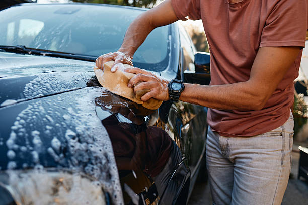 Adult man wearing casual clothes washing a black car hood with a sponge outdoors on a sunny day, creating soapy water suds. Focus on hands and car surface with natural lighting. Casual car cleaning activity.