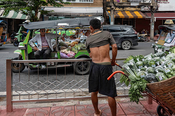 Bangkok, Thailand, October 1, 2025: A worker with a woven wicker cart piled with leafy greens on a street market stops and talks to a tuk-tuk driver who is also transporting vegetables.