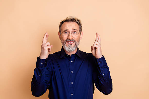 istock Mature man in formal button-up shirt with cheerful expression gesturing luck on beige background 2239741279