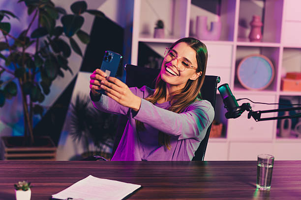 istock Young woman streaming using smartphone and microphone at modern home office with vibrant neon lighting in evening 2239743529