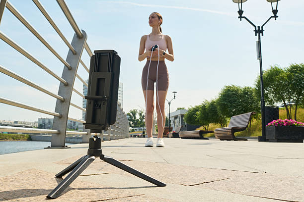 Young woman exercising outdoors with jump rope while recording workout on smartphone mounted on tripod standing on paved riverside promenade near metal railing