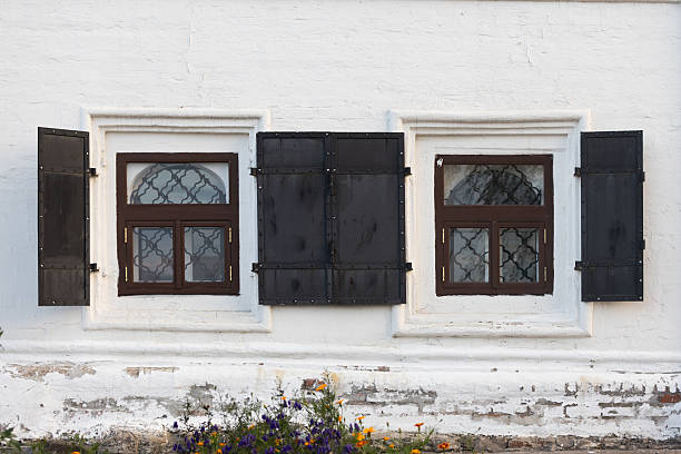 Square windows on ground floor with wooden frames and metal lattices and open black metal shutters on white stone house, Russia. Fragment of the facade of old historic house in Yeniseysk town, Russia