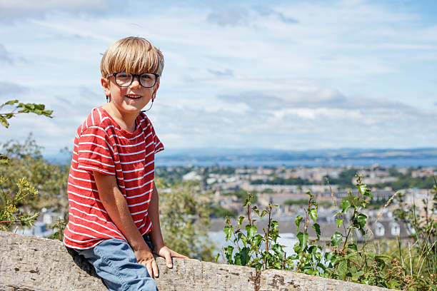 A young boy with glasses sits on a rock, smiling against a backdrop of a scenic view and clear skies in Calton Hill park, Edinburgh