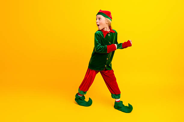 A cheerful child in a green and red elf costume walking on a yellow background, creating a whimsical holiday-inspired scene.