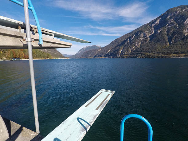 A diving board extends over the deep blue waters of Lake Achensee, offering a thrilling alpine perspective against the backdrop of the rugged mountains