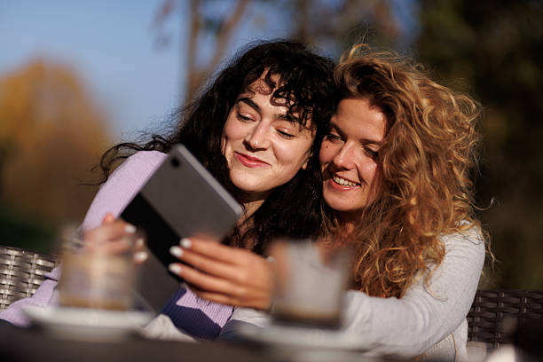 Two friends enjoying a sunny afternoon together while sharing laughter and a tablet in a cozy outdoor setting surrounded by greenery