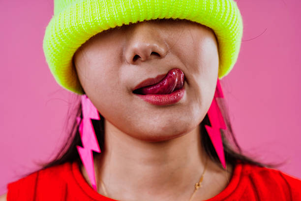 Young woman wearing a bright neon yellow beanie and vibrant pink lightning bolt earrings, showing a playful facial expression with her tongue sticking out on a pink background