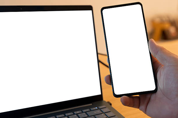 Close up of hand holding smartphone with blank white screen next to laptop on wooden desk. Modern technology background concept for mockup, digital device, business communication, creative workspace.