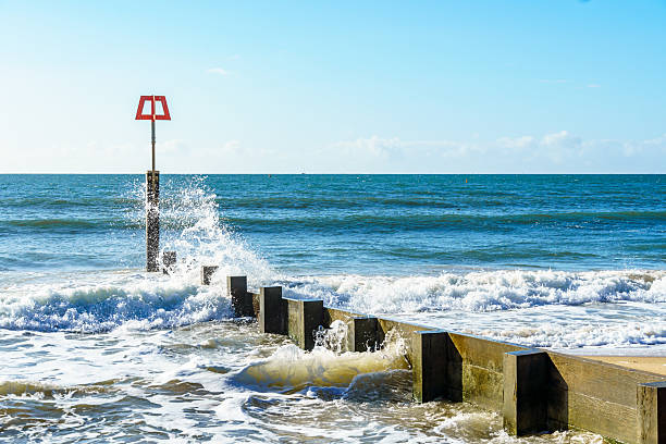 Dynamic ocean waves crashing against weathered wooden posts, creating a spray of water droplets under natural sunlight.