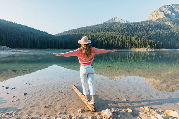 Young woman in hat and pink sweater balances on a log at the edge of a clear mountain lake. Scenic outdoor nature scene with forest and rocky peaks. Peaceful lakeside adventure.