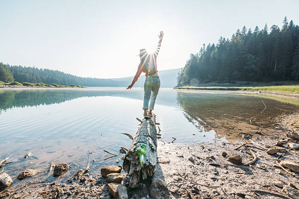 Young woman in hat and sweater balancing on a fallen log over a clear lake at sunrise. Outdoor adventure with serene water and forest background. Nature and mindfulness concept.