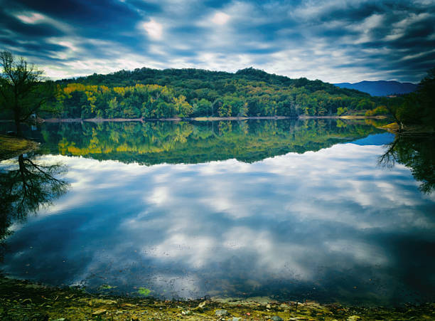 Calm lake mirror reflects vibrant forested hillside and cloudy sky, creating a serene, symmetrical landscape of clear water, autumn trees and peaceful wilderness scenery
