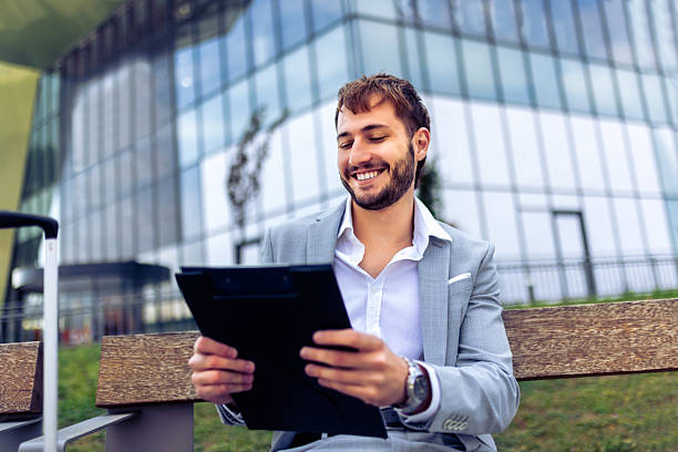 istock Businessman sitting on bench outdoors with suitcase reviewing clipboard 2239790401