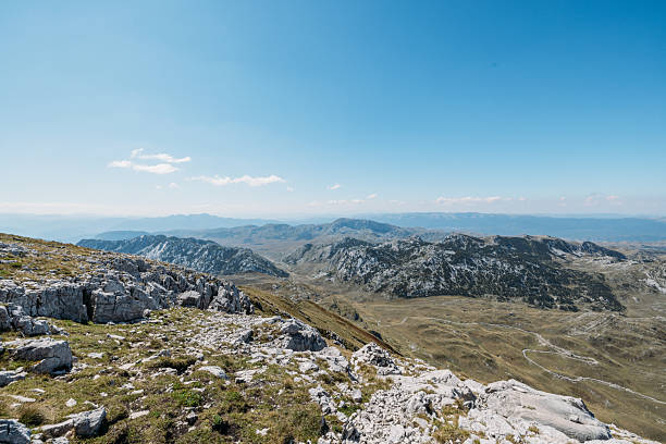 Expansive mountain ridge with rocky and grassy terrain under clear blue sky. Snowcapped peaks in the distance create a dramatic natural landscape. Scenic outdoor mountain environment.