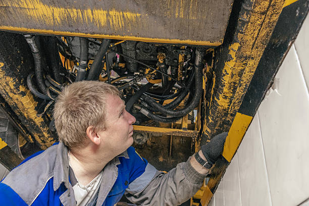A mechanic examines the inner workings of heavy machinery, focusing on maintenance and repair. The setting is industrial and technical, conveying a sense of diligence.