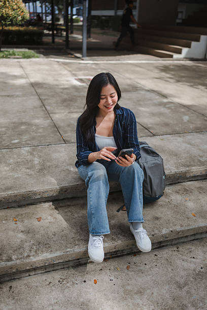 istock Young Asian woman sitting on concrete stairs on a campus, smiling while using her smartphone with a backpack next to her 2239797232