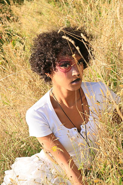 A waist up of a Black model sitting in tall dry grass on a sunny Summer day. She is wearing short black hair, makeup, a necklace and white t shirt.