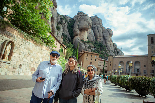 A family poses for a photo at the historic Montserrat monastery, with its dramatic rock formations towering in the background under a partly cloudy sky.