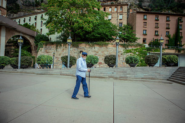 An older man strolls across a spacious plaza at Montserrat, holding his cane, with historic stone buildings and lush greenery behind him.