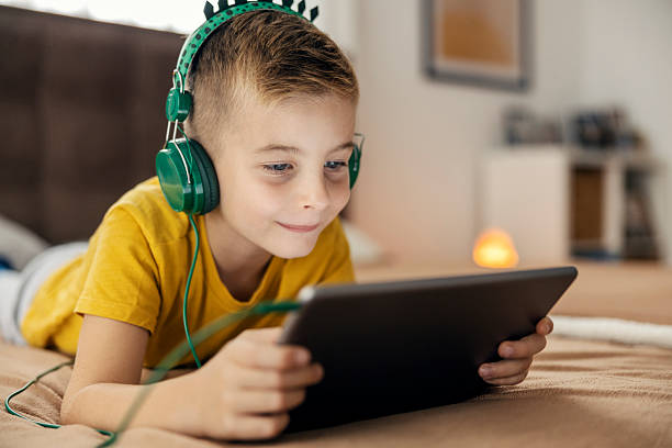 istock Young boy watching tablet with headphones lying on bed 2239814575