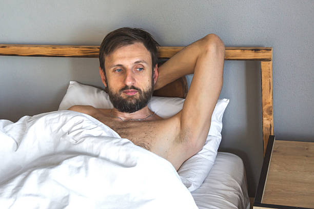 Man lying in bed with pensive expression, hand behind head, reflecting silently in minimalist bedroom, concept of midlife crisis and self-reflection