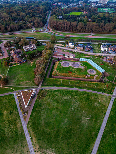 istock Aerial view of semi rural zone angular building with rooftop circles and green roofed triangle structure amid grassy fields, roads, forest edge, and sports field near clustered homes. 2239822968