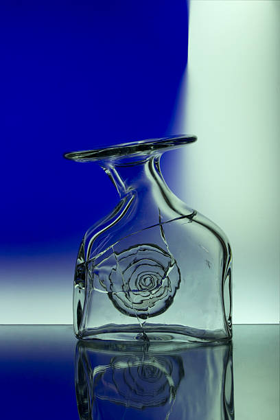 A close-up of a cracked transparent glass vase with abstract distortion and reflection on a glossy surface. The background features a gradient from deep blue to white, creating a modern, minimalist composition. Concept of fragility, imperfection, and beauty in broken objects.
