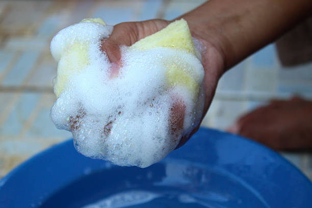 A pair of hands scrubs a green sponge in a bright green bucket filled with soapy water. The scene conveys cleaning, domestic chores, and fresh, hygienic home care.