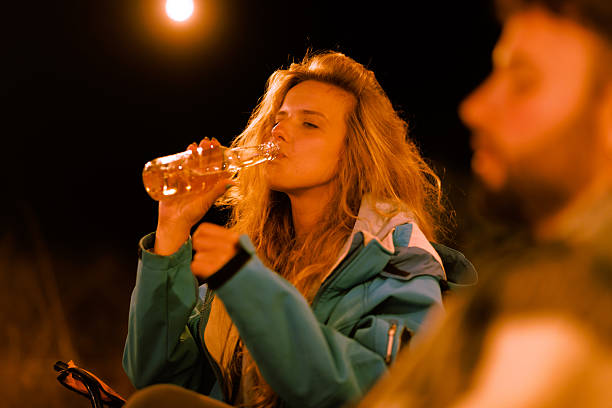 A young woman relaxes by a campfire at night, drinking from a bottle while sharing laughter with friends. The warm light creates a cozy atmosphere, perfect for bonding moments.