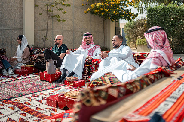 A diverse group of business people, including men and women, engage in relaxed conversations in a traditional Arabian Majlis. The setting is enhanced by colorful patterns and outdoor surroundings.