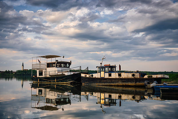 Old river ships under the Serbian flag, standing on a pier on the Danube against a cloudy sky and an old bell tower.