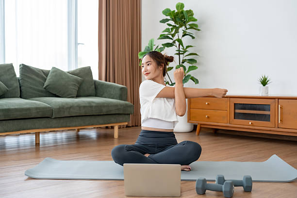 istock Young Asian woman stretching yoga workout on exercise mat while online training class with computer laptop in living room 2239858589
