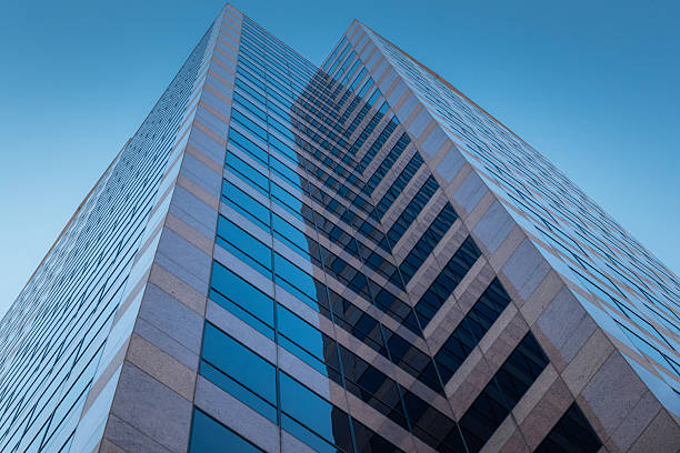 High-rise glass building with angular design featuring blue and gray tones. Architectural perspective captured from low angle showcasing symmetrical lines and modern construction techniques against clear blue sky background.