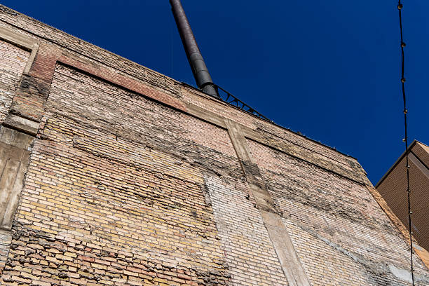 Aged industrial building exterior with textured brick surface, exposed vertical pipe, and clear blue sky background. Architectural details showcase layers of construction materials and aging process of urban infrastructure.
