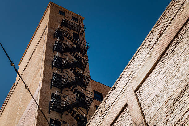 Exterior view of multi-story brick building with metal fire escape staircase zigzagging vertically against clear blue sky. Architectural details highlight industrial urban construction with weathered brick wall texture and angular metal stairs.